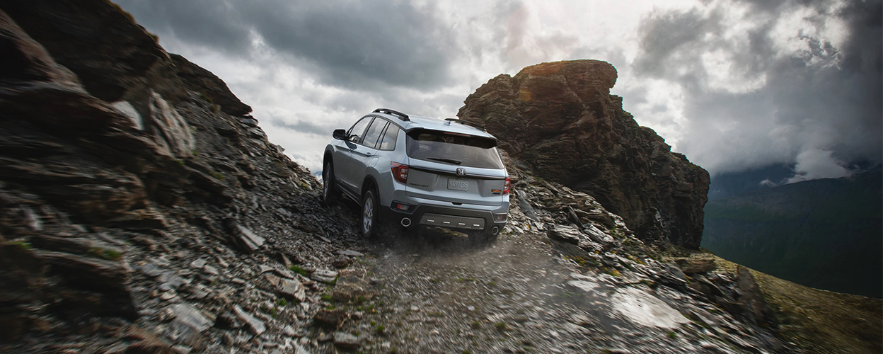 3/4 rear view of grey Honda Passport driving up dirt road.