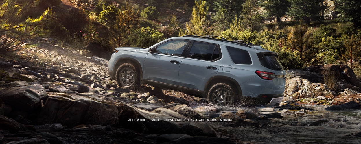 3/4 side view of a blueish grey Pilot driving through a creek and onto an incline on in a temperate rainforest.