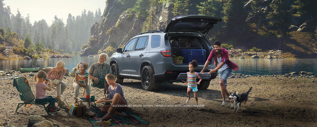 3/4 rear view of blueish grey Pilot with tailgate open parked by a river in a temperate rainforest. Three generations of family are around it enjoying a picnic.