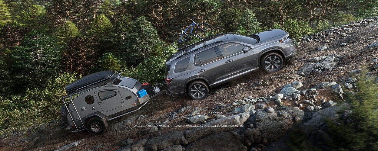 Wide sideview of grey Pilot, with a bike on the roof rack, towing a grey trailer uphill on a dirt road in a temperate rainforest.