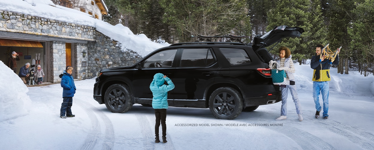 Plan latéral de trois quarts d’un Pilot TrailSport noir stationné dans l’entrée enneigée d’un chalet moderne avec la porte de garage ouverte. Trois générations d’une même famille l’entourent. Maman et papa déchargent le hayon ouvert.