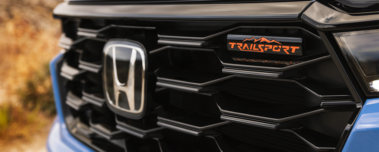 Closeup of front grille on light blue Pilot, showcasing the Honda logo and the TrailSport emblem. 