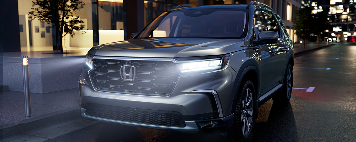 Plan de trois quarts avant d’un Pilot gris stationné en bordure de rue en ville avec phares avant allumés. 