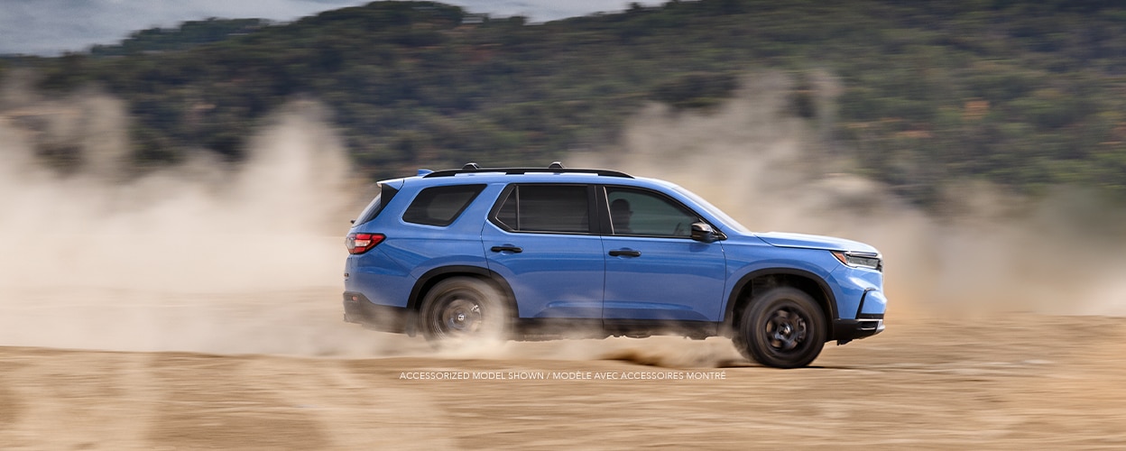 Wide sideview of light blue Pilot driving on a desert plain kicking up dust.