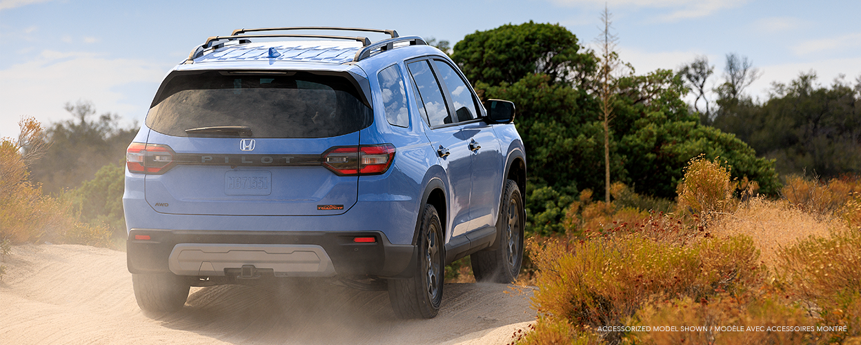 3/4 rear view of light blue Honda Pilot driving on a desert dirt road amid desert plants.