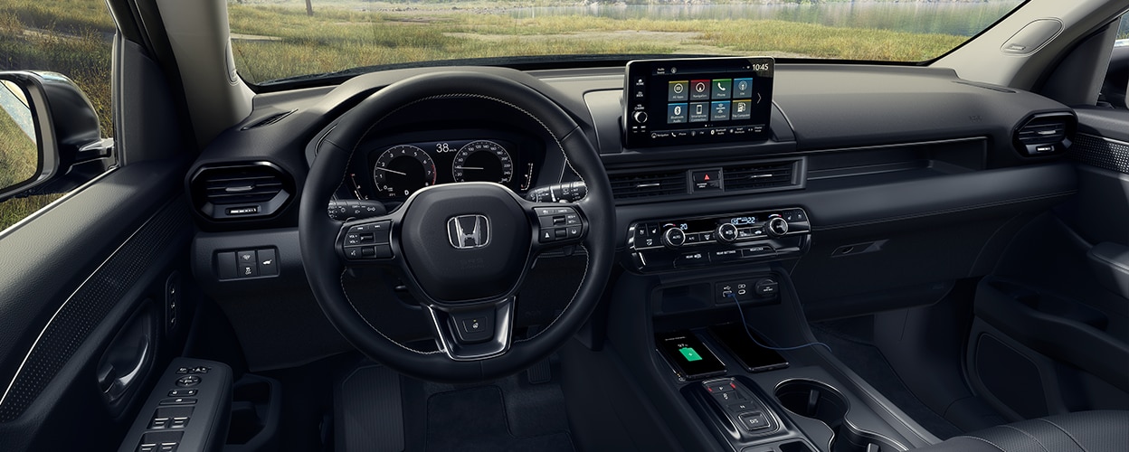 Panoramic wide view of steering wheel, dashboard, and centre console. Out the windshield we see a beautiful forest vista. 