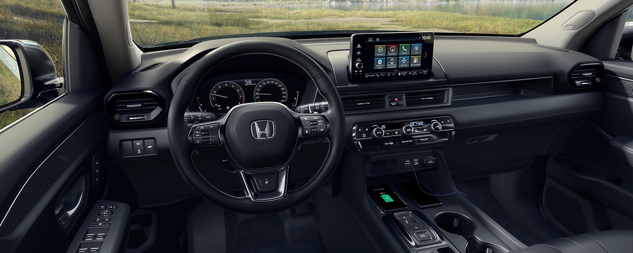 Panoramic wide view of steering wheel, dashboard, and centre console. Out the windshield we see a beautiful forest vista.