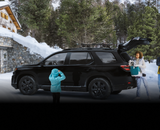 Plan latéral de trois quarts d’un Pilot TrailSport noir stationné dans l’entrée enneigée d’un chalet moderne avec la porte de garage ouverte. Trois générations d’une même famille l’entourent. Maman et papa déchargent le hayon ouvert.