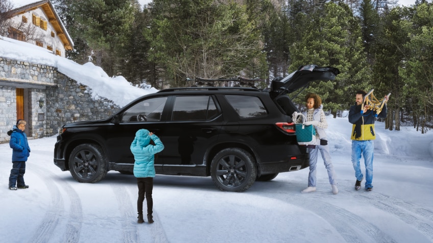 Plan latéral de trois quarts d’un Pilot TrailSport noir stationné dans l’entrée enneigée d’un chalet moderne avec la porte de garage ouverte. Trois générations d’une même famille l’entourent. Maman et papa déchargent le hayon ouvert.