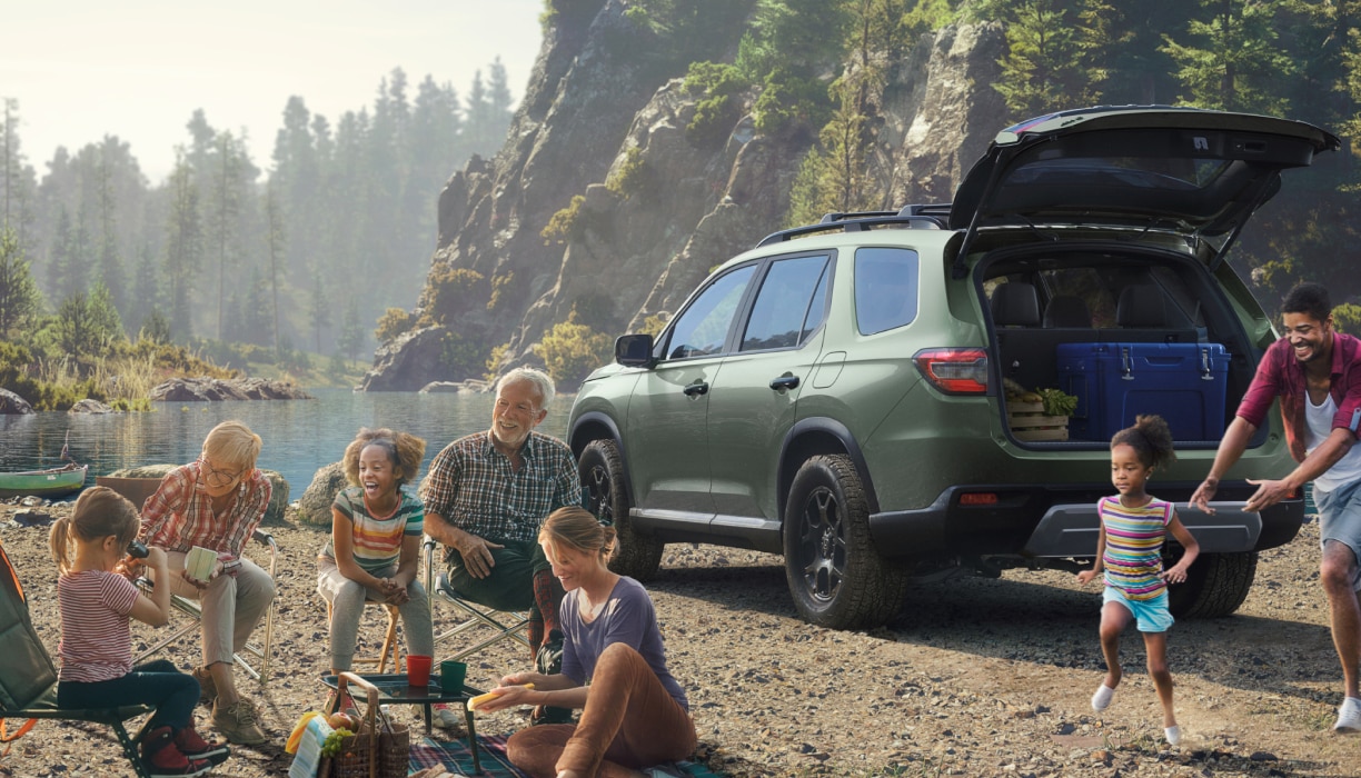 Vue de trois quarts arrière d’un Pilot vert garé près d’une montagne au bord d’un lac. Le hayon est ouvert, révélant une boîte bleue et une caisse en bois à l’intérieur. À proximité, un parent joue avec une petite fille, tandis qu’une famille est assise sur des chaises pliantes autour d’une couverture de pique-nique.