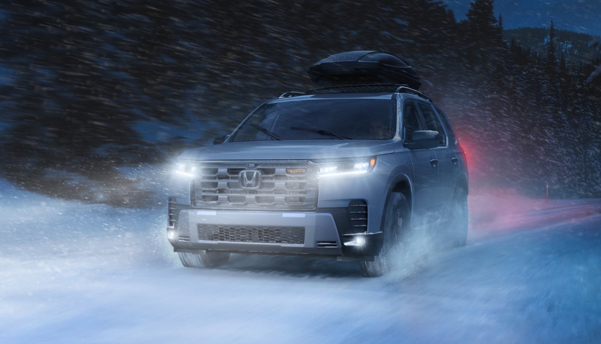Front view of a silver Pilot driving on a dark, snow-covered road with headlights on. Blowing snow surrounds the vehicle and a cargo box is mounted on the roof.