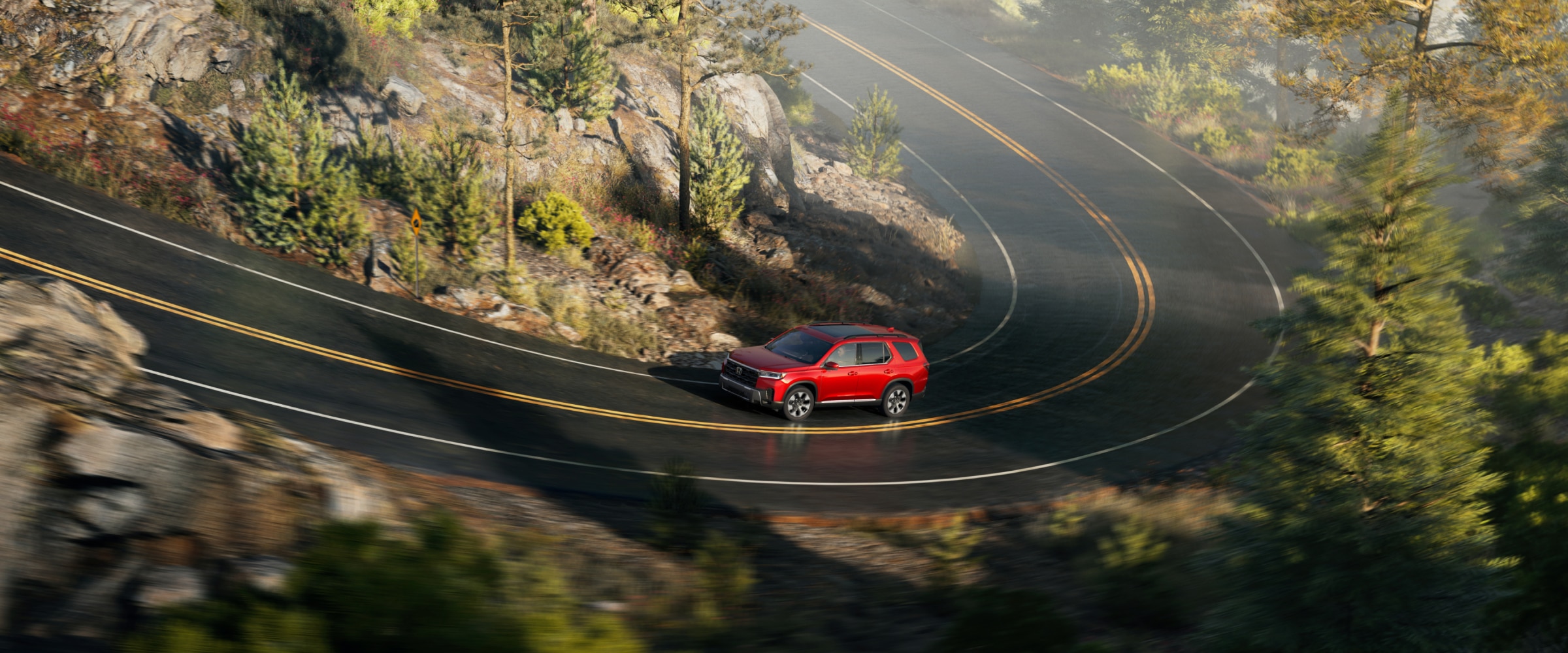 Side view of a red Pilot driving along a winding highway in a rugged landscape of rocks, bushes, and trees.