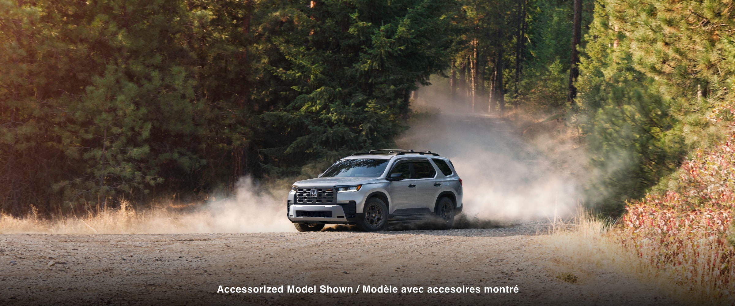 3/4 front side view of a silver Pilot driving on a dirt road through a forest, with dust trailing behind its wheels.