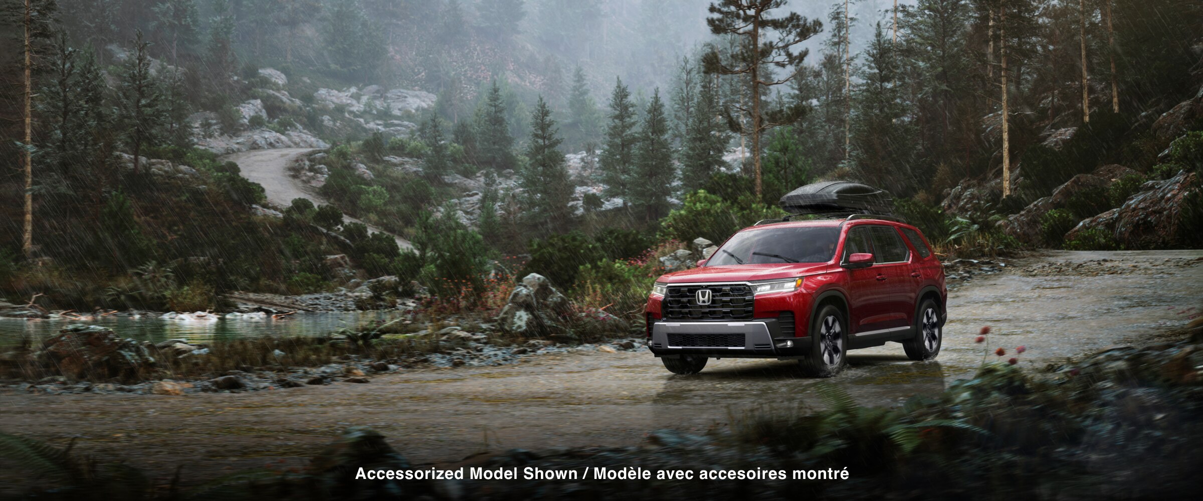 Vue de trois quarts avant d’un Pilot rouge roulant sur un chemin de terre à travers une forêt sous la pluie, avec une caisse de chargement montée sur le toit. 