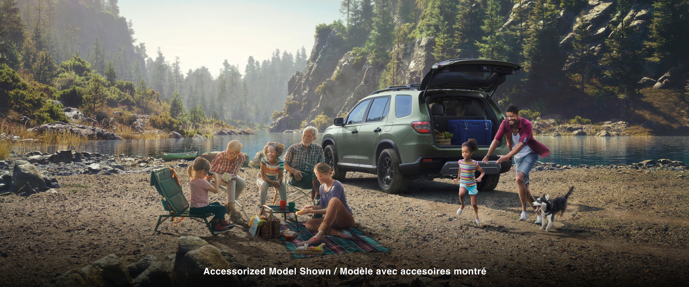 3/4 rear view of a green Pilot parked near a lakeside mountain. The tailgate is open, revealing a blue box and wooden crate inside. Nearby, a parent chases a young girl, while a family sits on folding chairs around a picnic blanket.