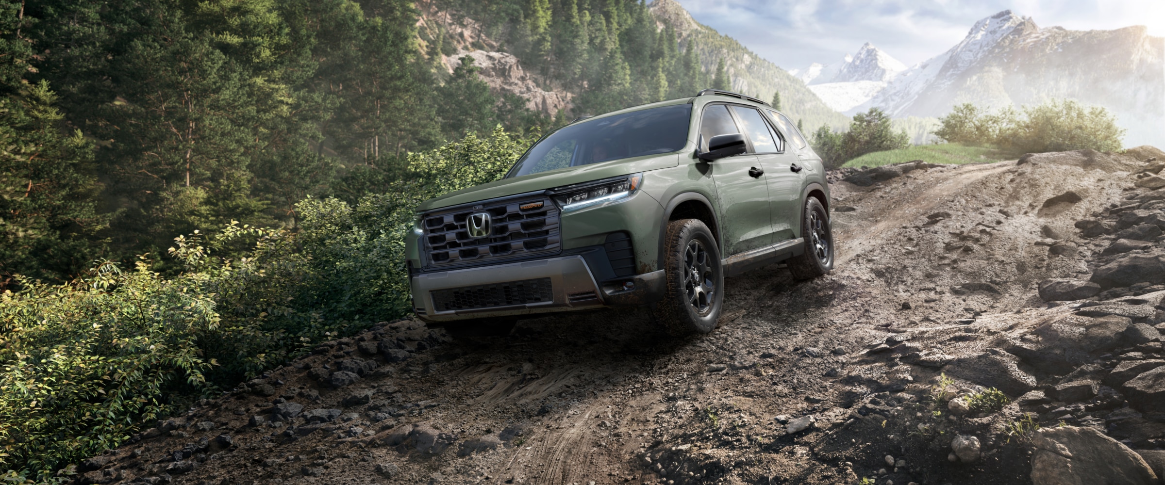 Vue de trois quarts avant d’un Pilot vert descendant un chemin de terre dans la forêt, avec des montagnes lointaines en arrière-plan.