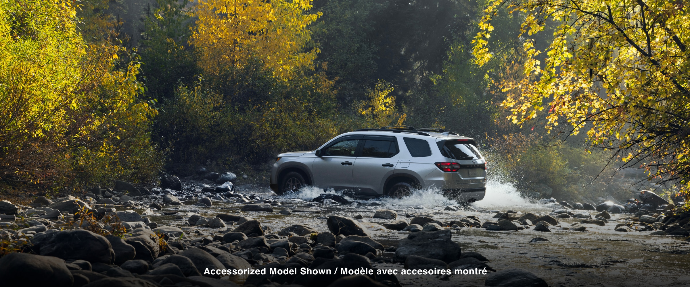 Side view of a silver Pilot driving across a shallow, rocky riverbed in a forest, water splashing around its tires.