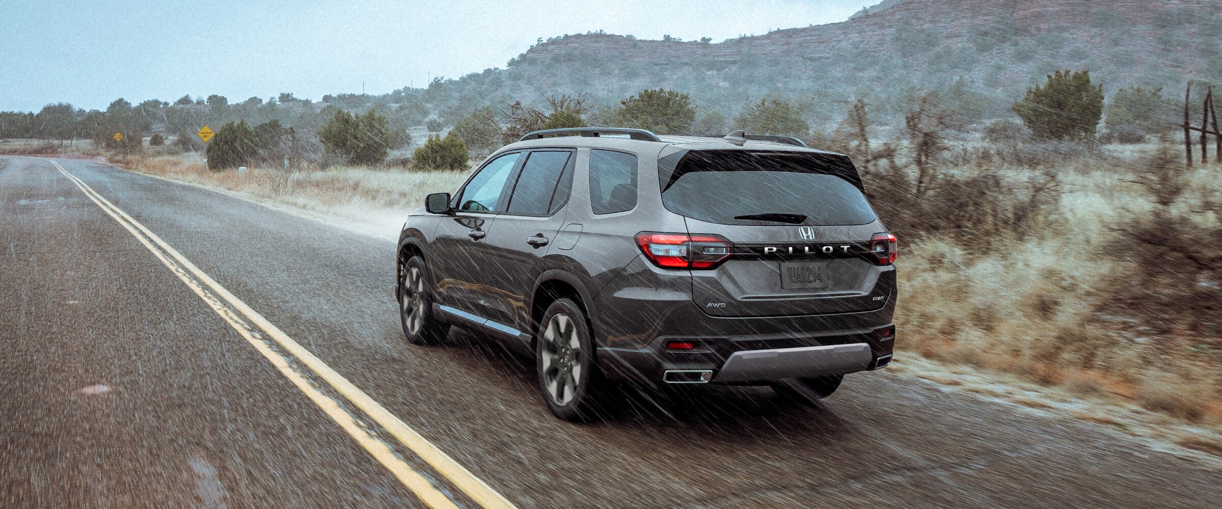 Vue de trois quarts arrière d’un Pilot gris roulant sous la pluie sur une autoroute dans un paysage accidenté de plantes et de broussailles.