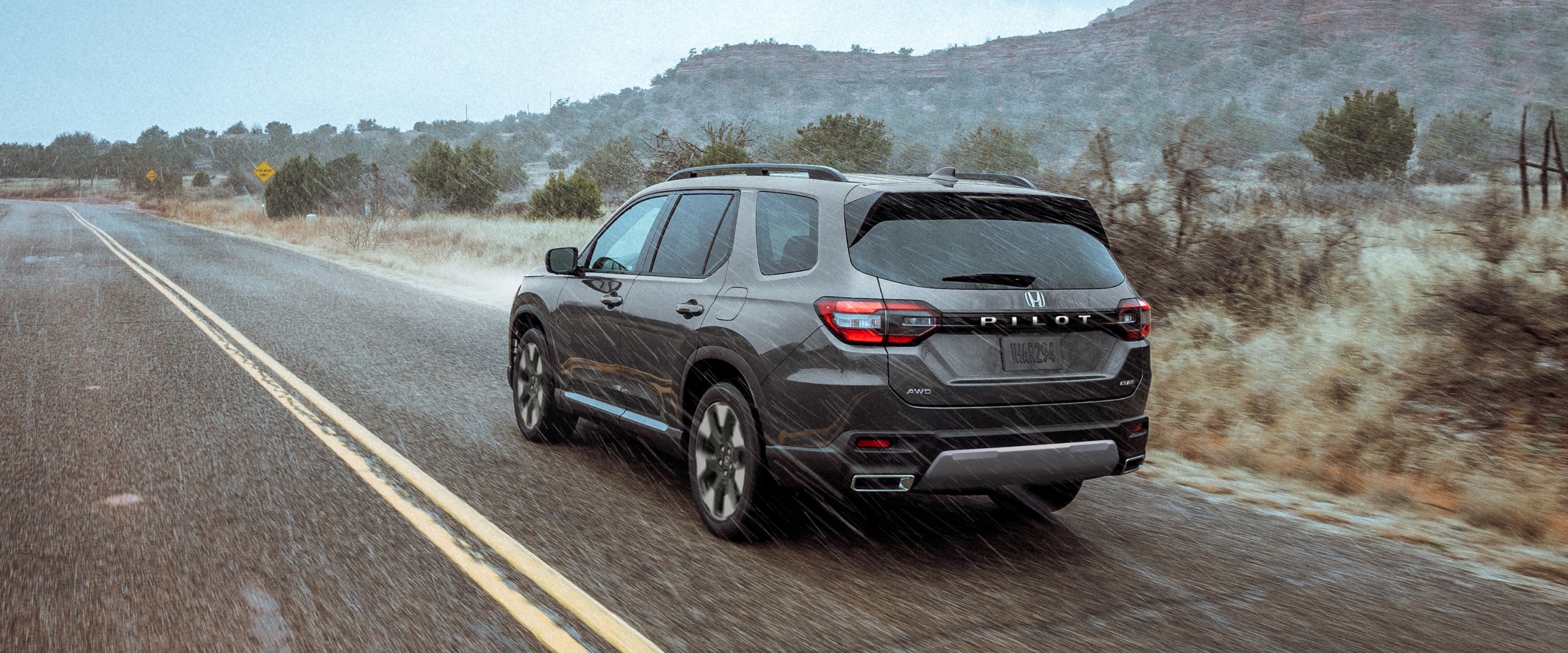 3/4 rear view of a grey Pilot driving through the rain on a highway set against a rugged landscape of plants and brush.