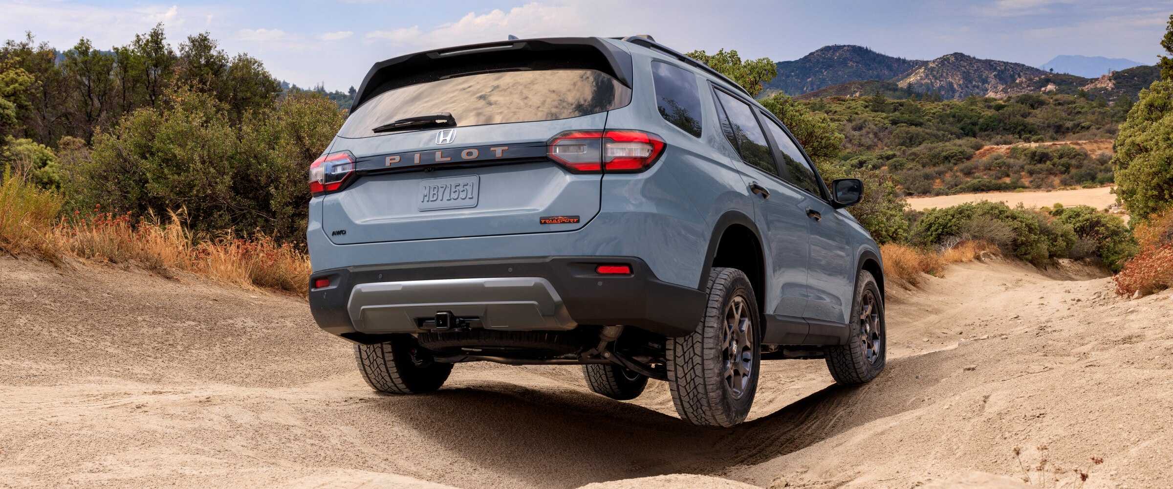 Vue de trois quarts arrière d’un Pilot bleu clair roulant sur un terrain accidenté avec des montagnes en arrière-plan. 