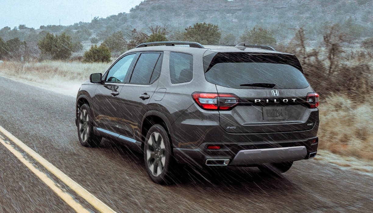 3/4 rear view of a grey Pilot driving through the rain on a highway set against a rugged landscape of plants and brush.