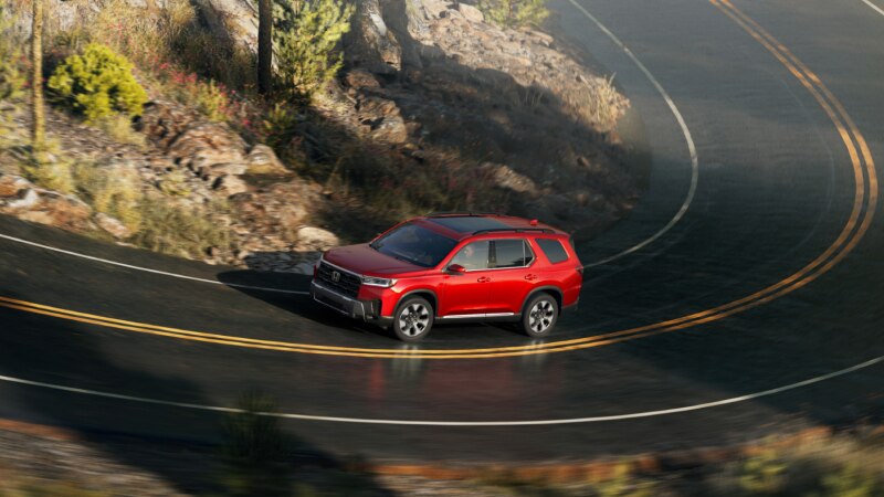 Vue latérale d’un Pilot rouge roulant sur une autoroute sinueuse dans un paysage accidenté de rochers, de buissons et d’arbres.
