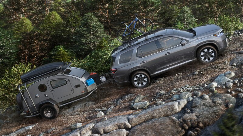 Vue latérale d’un Pilot gris gravissant une pente en terrain rocailleux, avec un vélo fixé sur le toit et une remorque attelée à l’arrière.