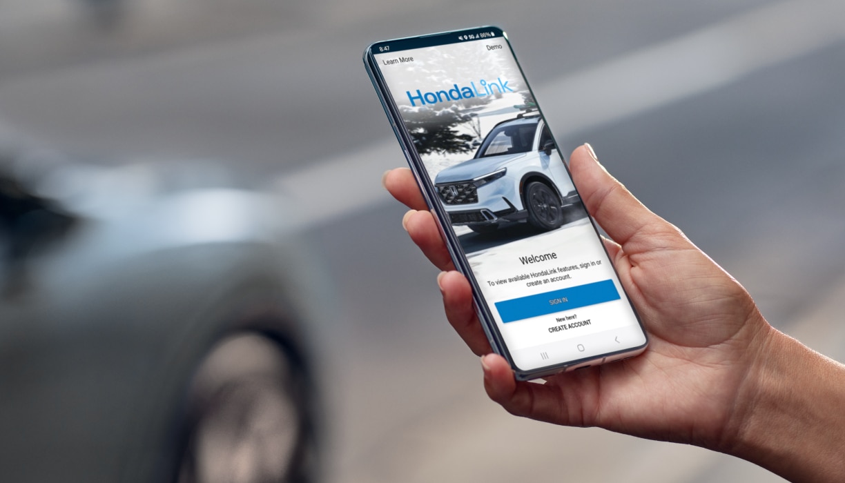 Gros plan d’une main utilisant l’application téléphonique HondaLink dans une rue. Véhicule gris garé à l’arrière-plan.