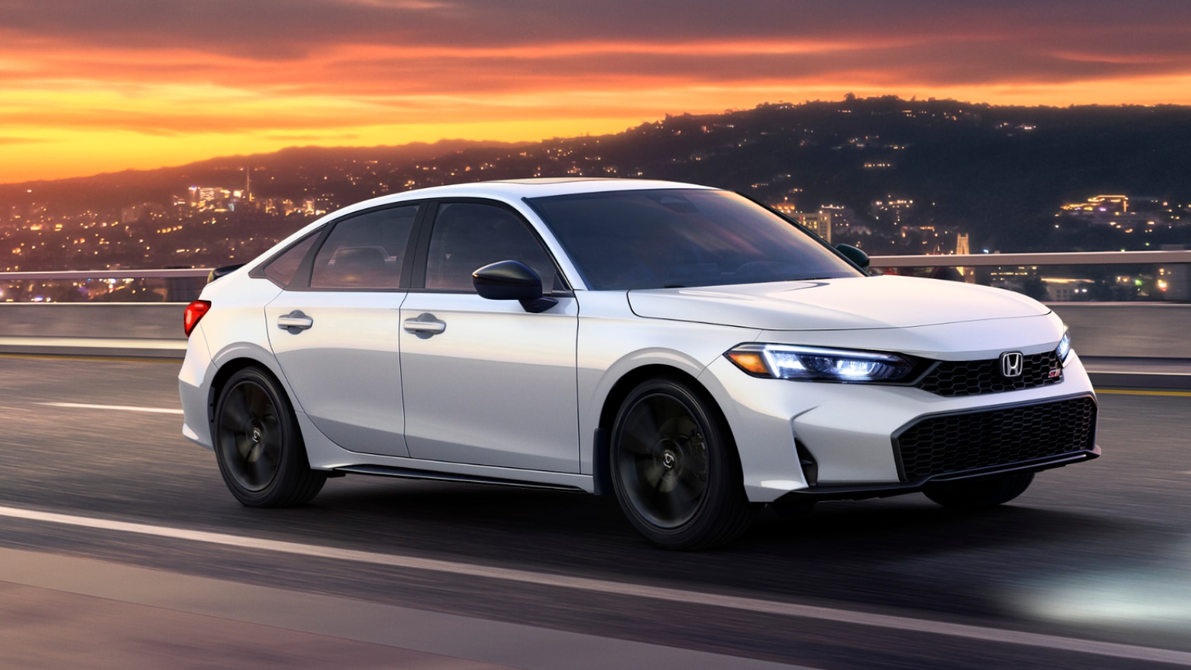 3/4 front side view of white Civic Si driving on a highway at sunset with city lights glowing on a hilly backdrop.