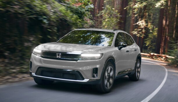 3/4 front view of white Prologue taking a turn on paved road in a temperate forest with the headlights on.