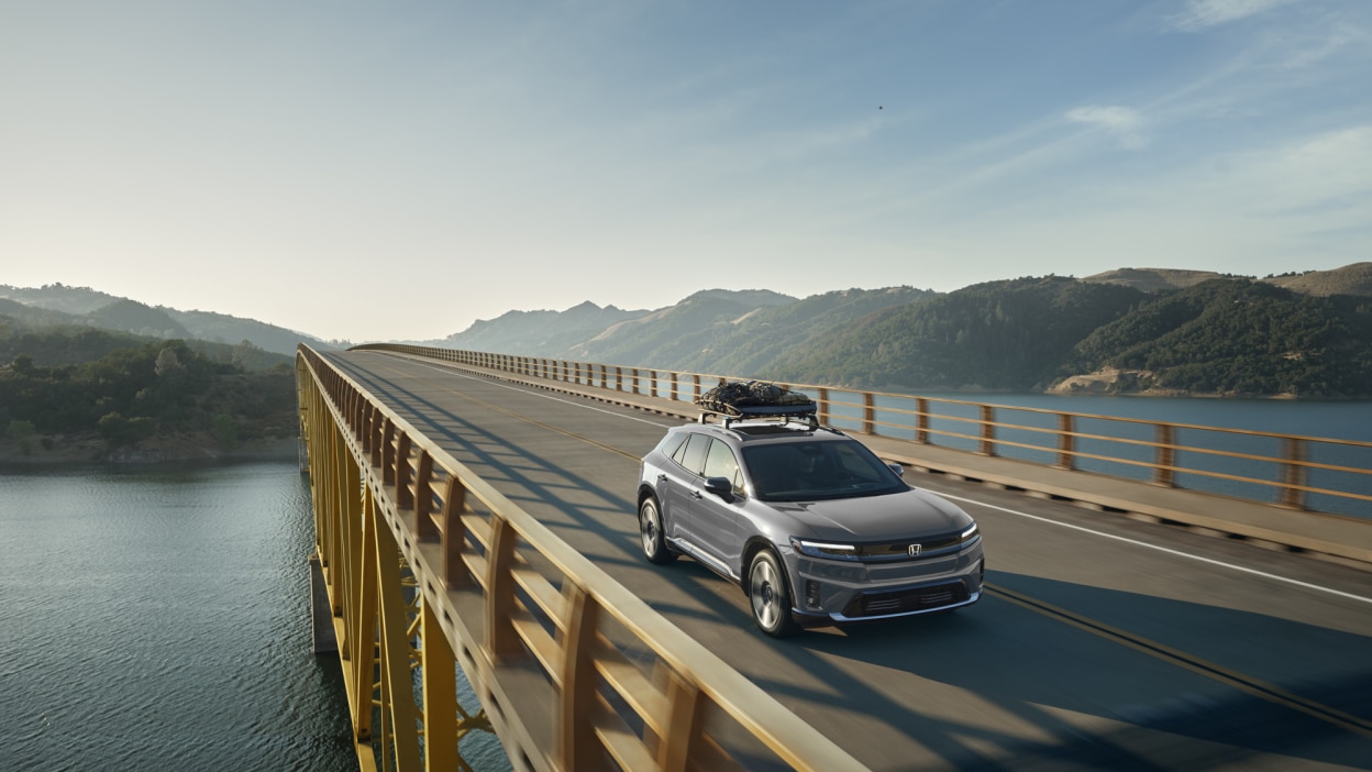 Wide 3/4 front view of grey Prologue with cargo on the roof rack driving over a bridge.