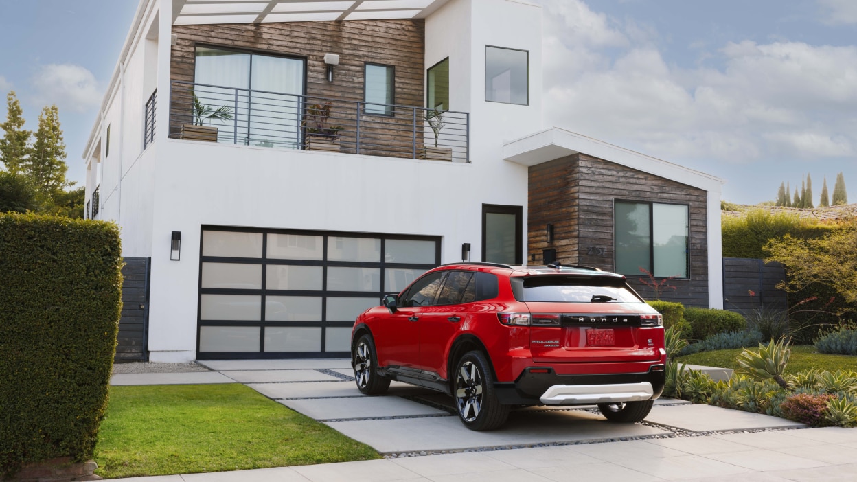 3/4 rear view of red Prologue parked in driveway in front of a white modern house with some wood siding.