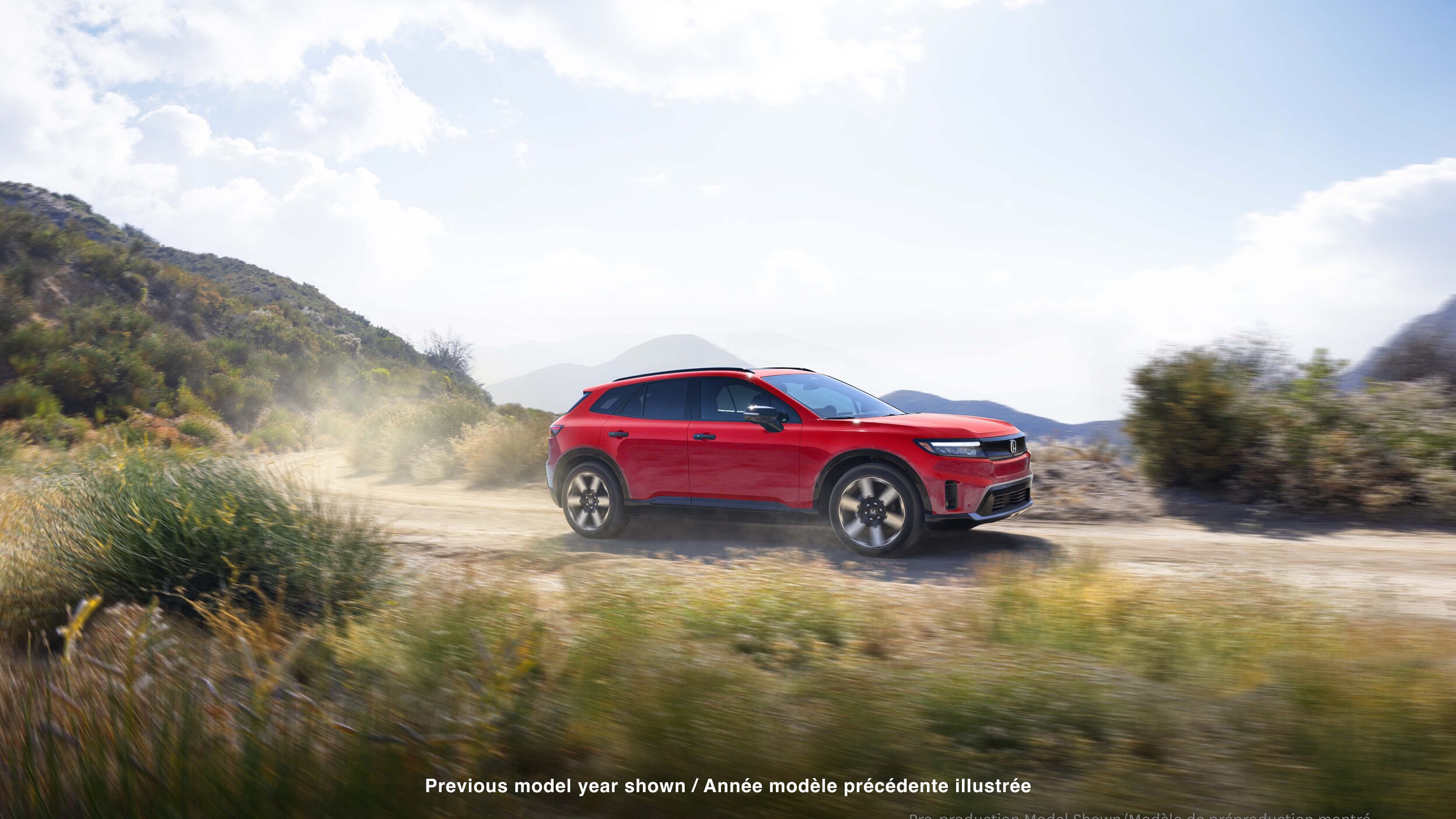 Wide 3/4 side view of red Prologue driving on desert road, kicking up dust. 