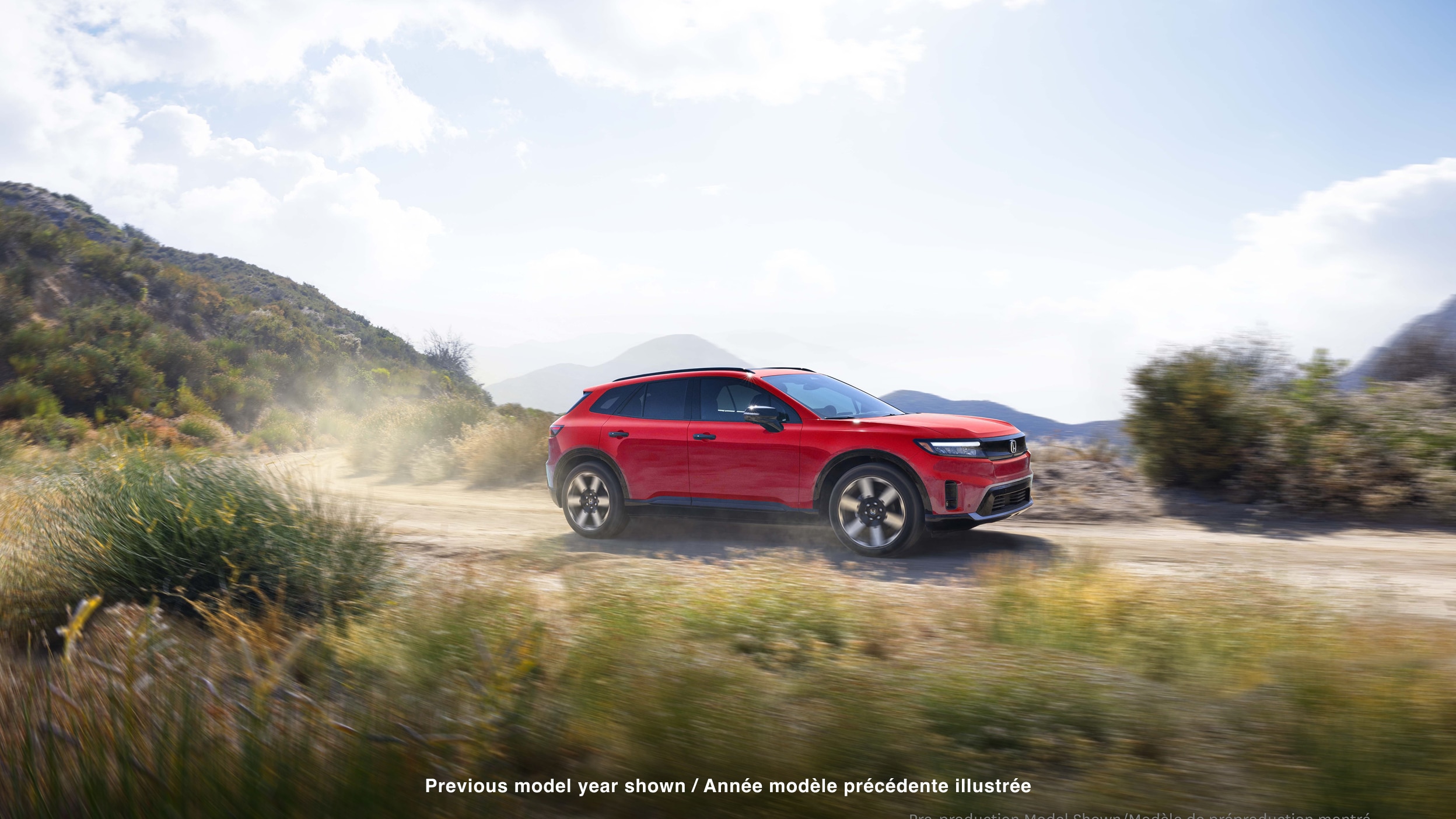 Wide 3/4 side view of red Prologue driving on desert road, kicking up dust.