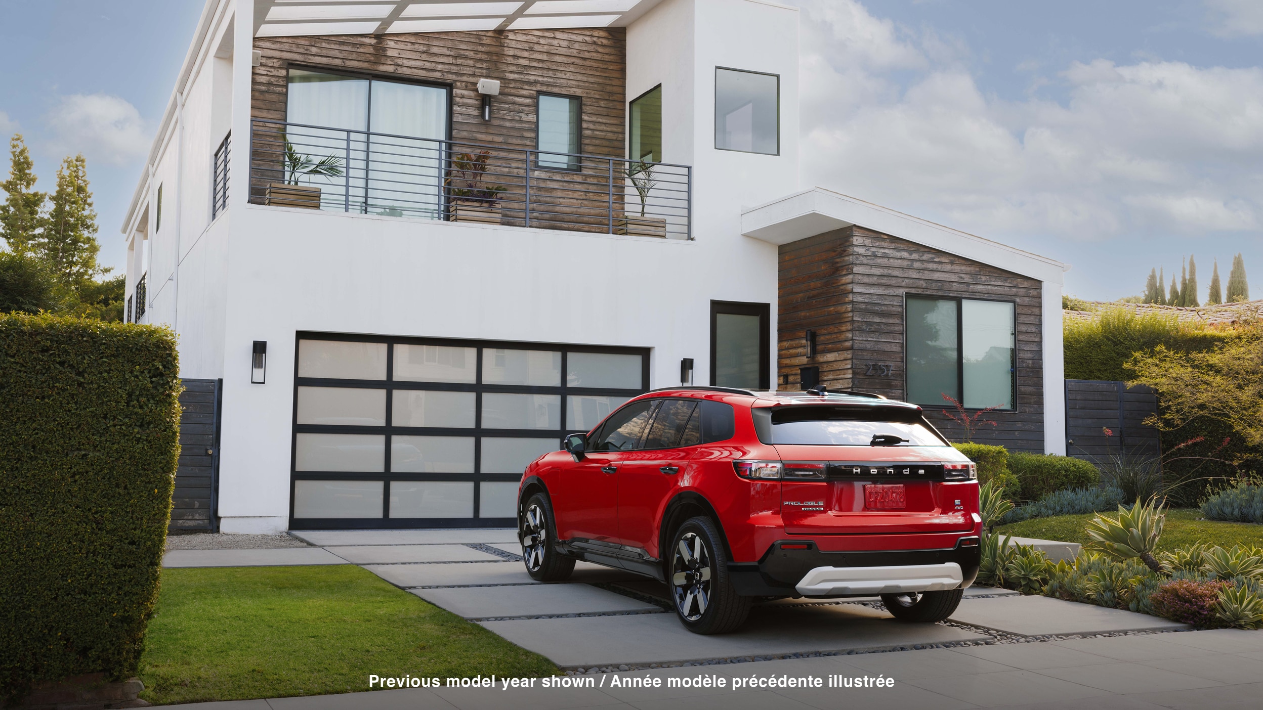 3/4 rear view of red Prologue parked in driveway in front of a white modern house with some wood siding.