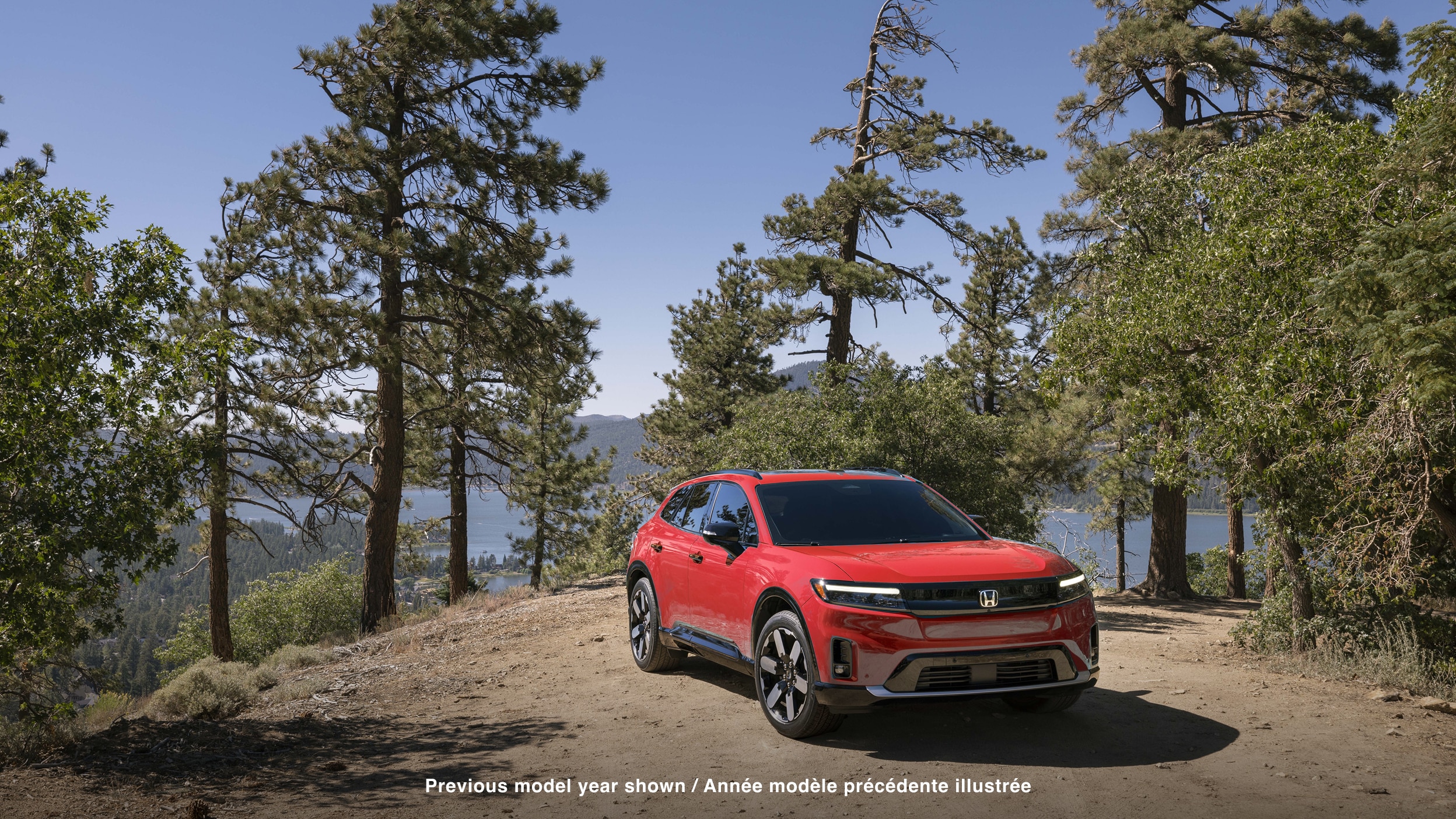 3/4 front view of red Prologue parked amongst trees in a forest. In the background we see a lake and mountians.