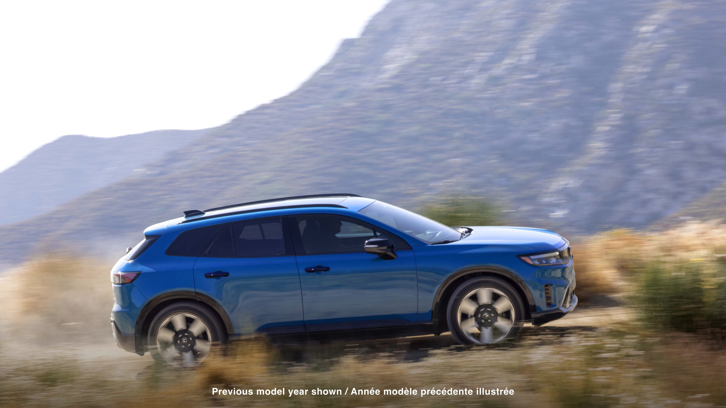 Wide side view of blue Prologue driving on desert road, kicking up dust. 