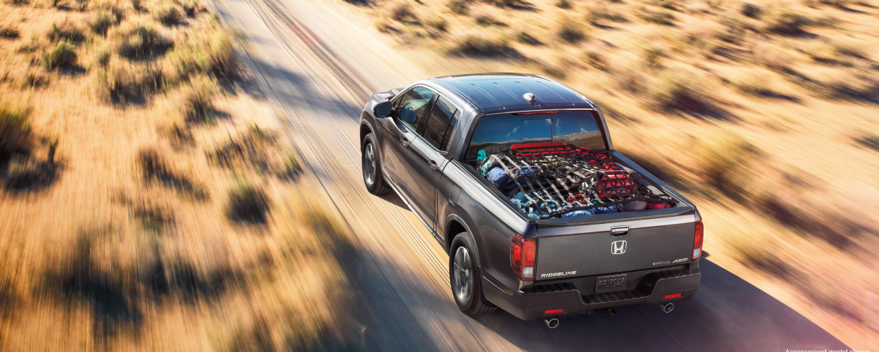 3/4 rear bird’s eye view of grey Honda Ridgeline driving on desert road. Truck-bed loaded with stuff.