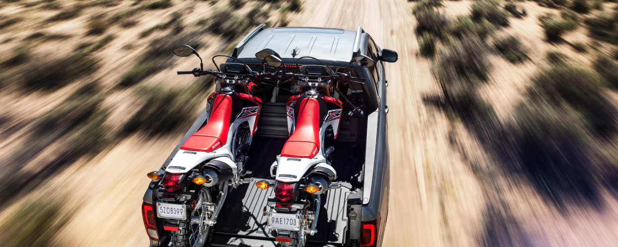 3/4 rear bird’s eye view of grey Ridgeline driving on desert road with two motocross bikes in truck-bed.