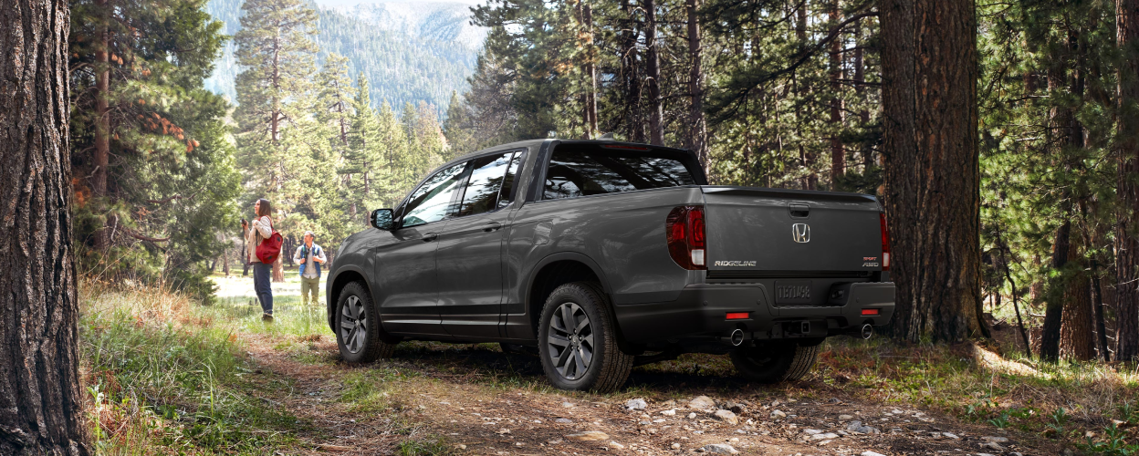 3/4 rear view of grey Ridgeline parked in forest. A couple standing in front it take in the scenery.