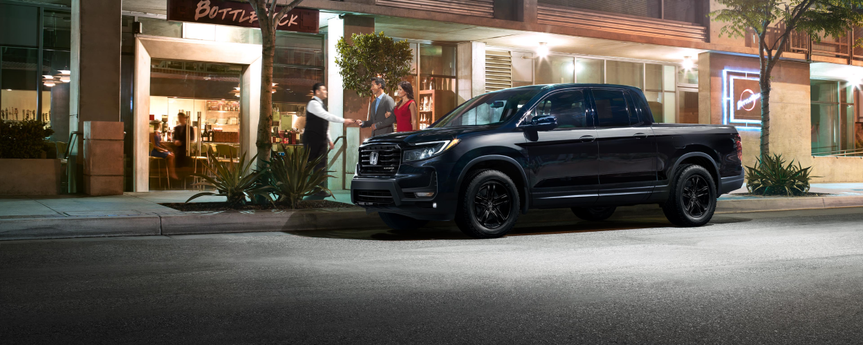 Side view of black Ridgeline parked curbside in city. A couple greets the host of the restaurant.