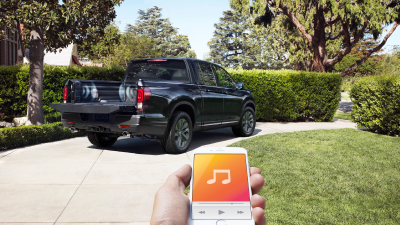 3/4 rear view of Ridgeline in driveway. Hand holding phone with music app open in foreground.