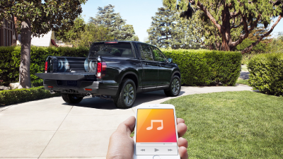 3/4 rear view of Honda Ridgeline in driveway. Hand holding phone with music app open in foreground.