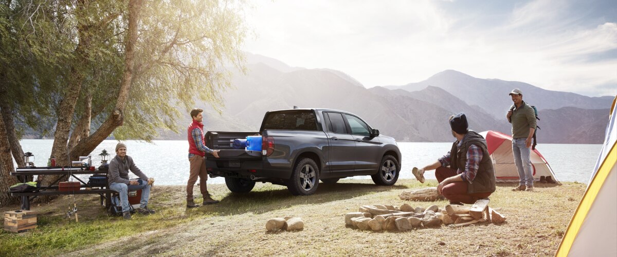 3/4 rear view of grey Ridgeline parked lakeside. A man unloads it, tailgate door open. His three buddies set up the campsite and campfire.