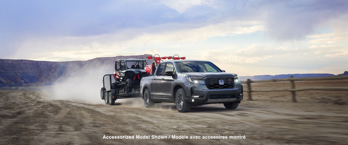 3/4 front wide view of grey Ridgeline driving in the desert with two motocross bikes in truck-bed, towing a trailer with a dune buggy.