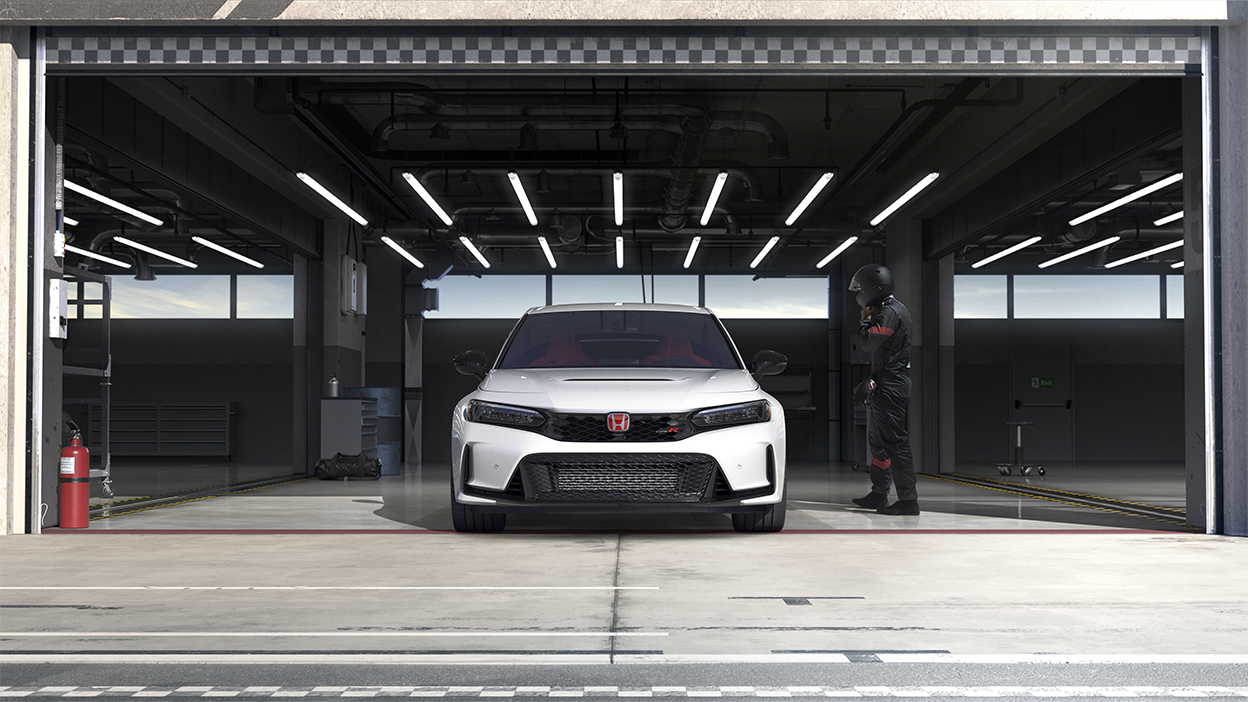 Front view of a white Type R parked in racing garage. A race car driver in black coveralls and helmet nears it. 
