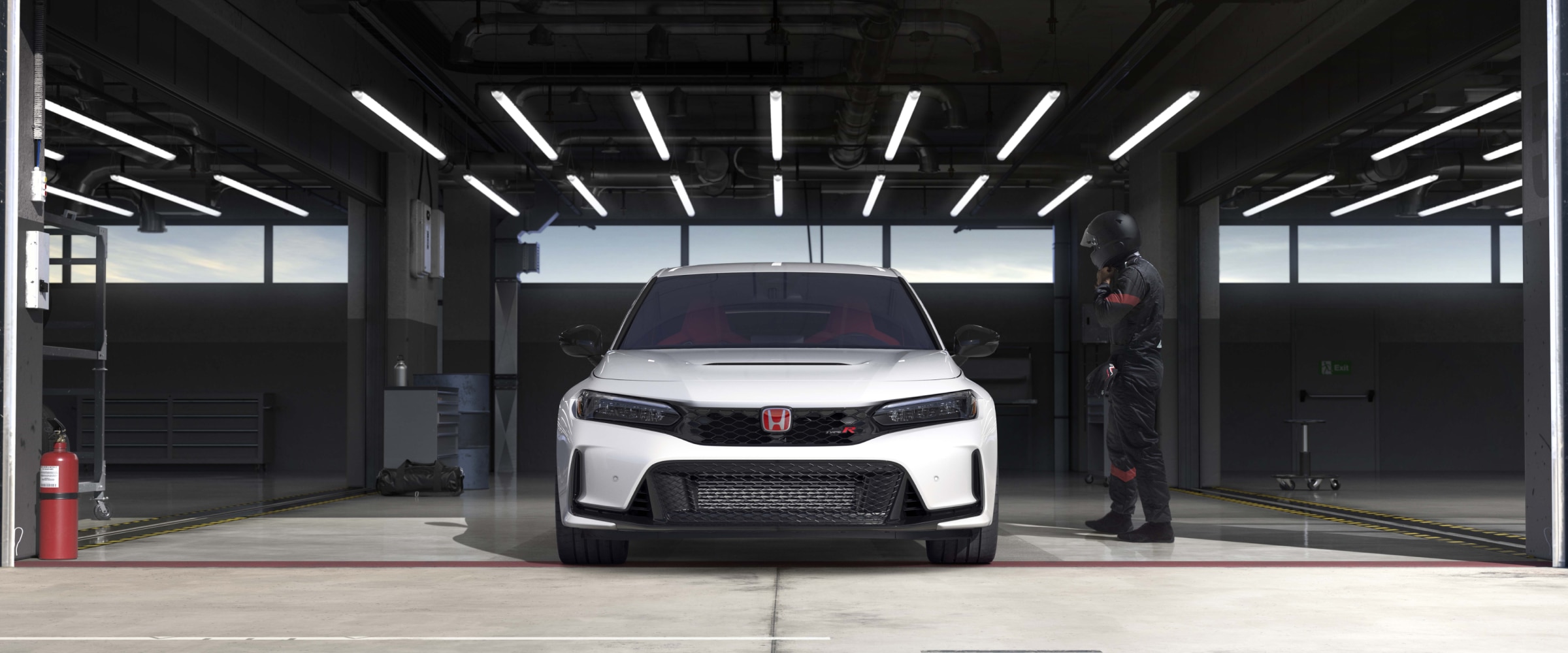 Front view of a white Type R parked in racing garage. A race car driver in black coveralls and helmet nears it.