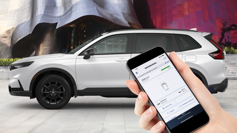 Side view of a white Honda vehicle with a hand holding a phone in the foreground. The Hondalink™ interface appears on the screen.