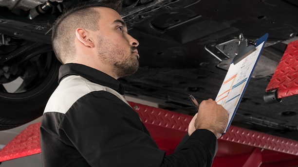 A Honda technician looking under a Honda vehicle and making notes on a clipboard. 
