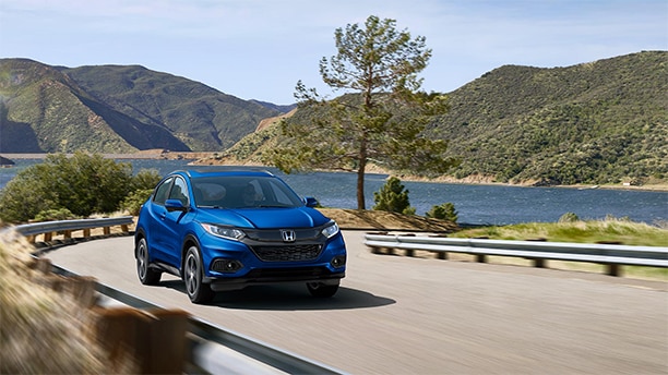 Low-angle view of a Honda HR-V in Aegean Blue Metallic driving on a curvy mountain road with a lake in the background.